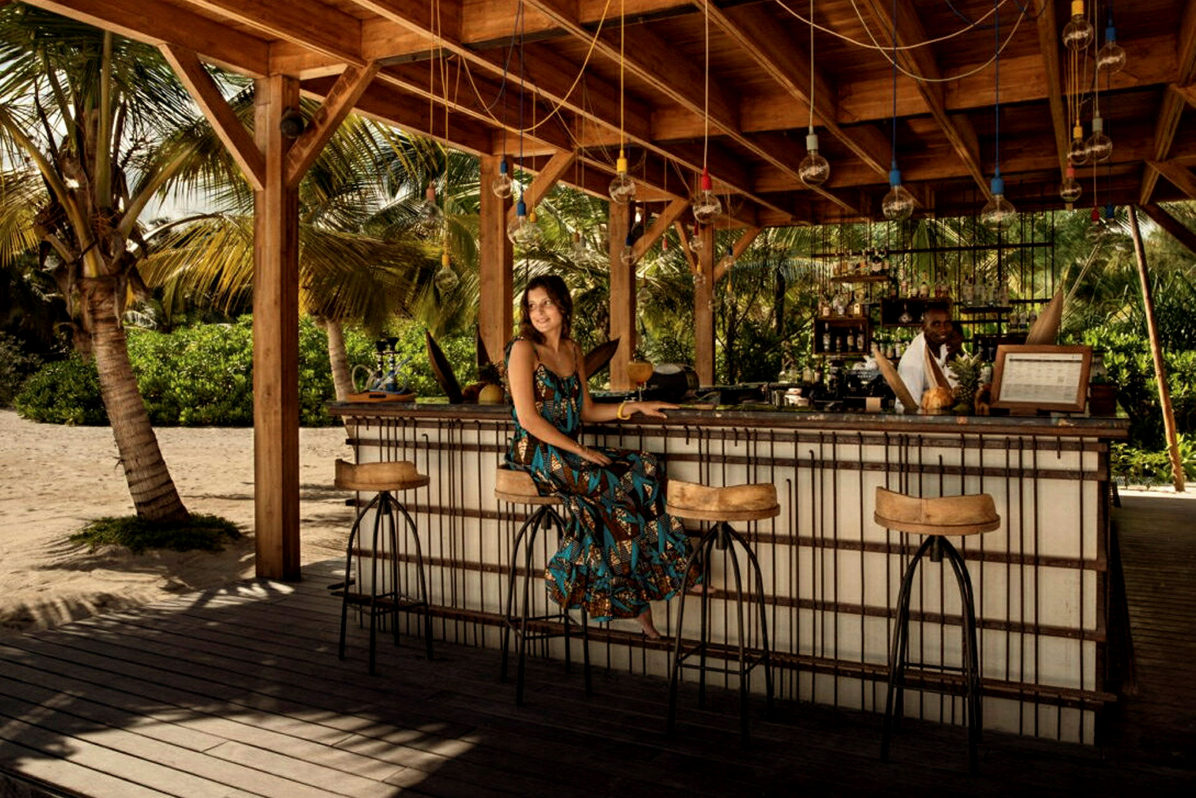 En strandbar under ett träskjul med höga pallar och palmträd i bakgrunden. (AI-genererad text)