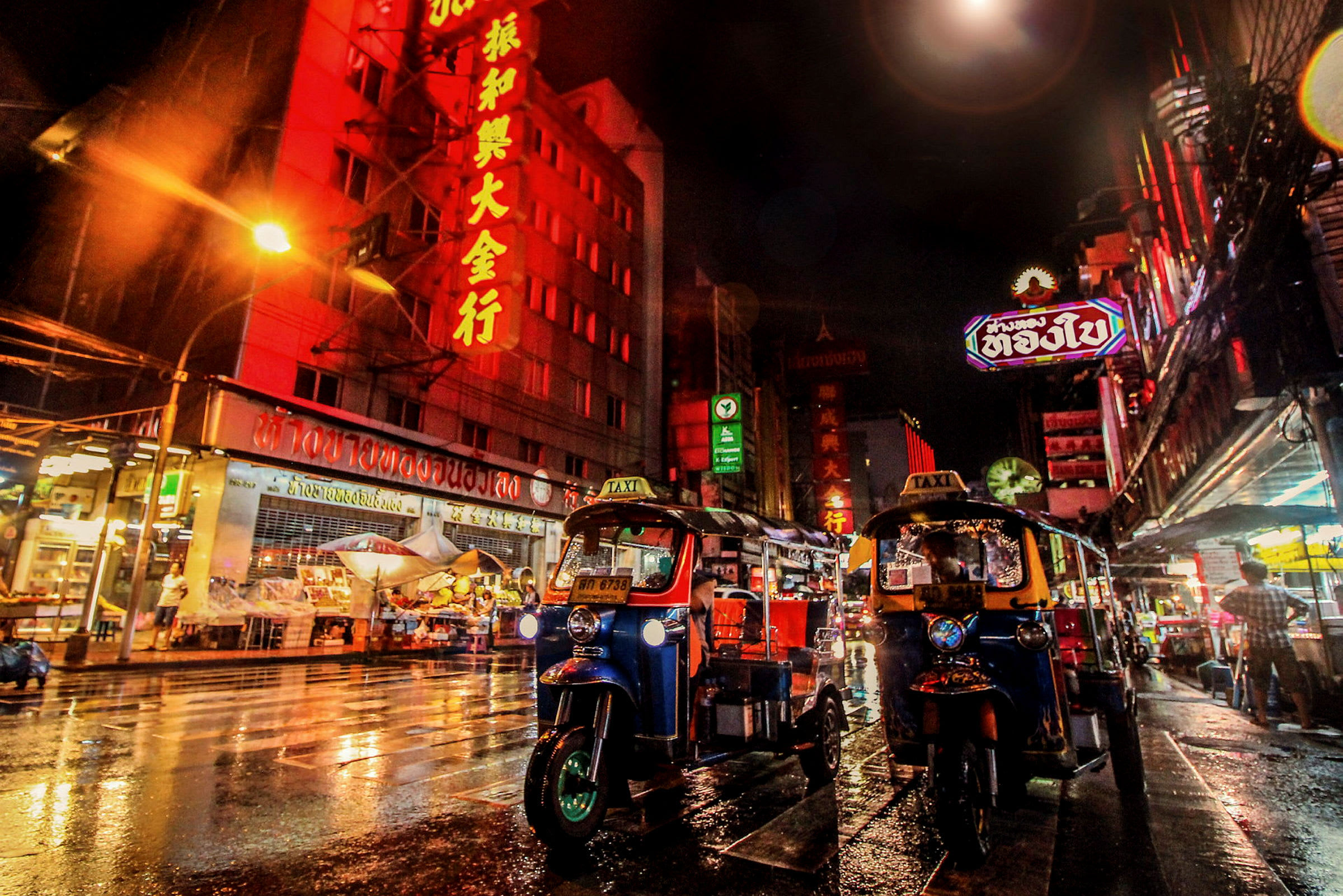 Tuk-tuk taxibilar på en livlig, regnvåt stadsgata på natten, omgiven av färgglada byggnader med neonljus. (AI-genererad text)
