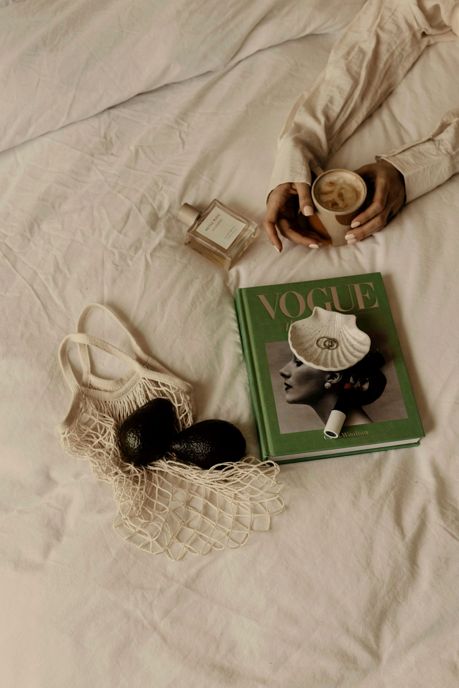 Bilden visar en bok med texten 'VOGUE' på omslaget, två avokador i en nätpåse, en parfymflaska, och en person som håller en kopp kaffe. (AI-genererad text)