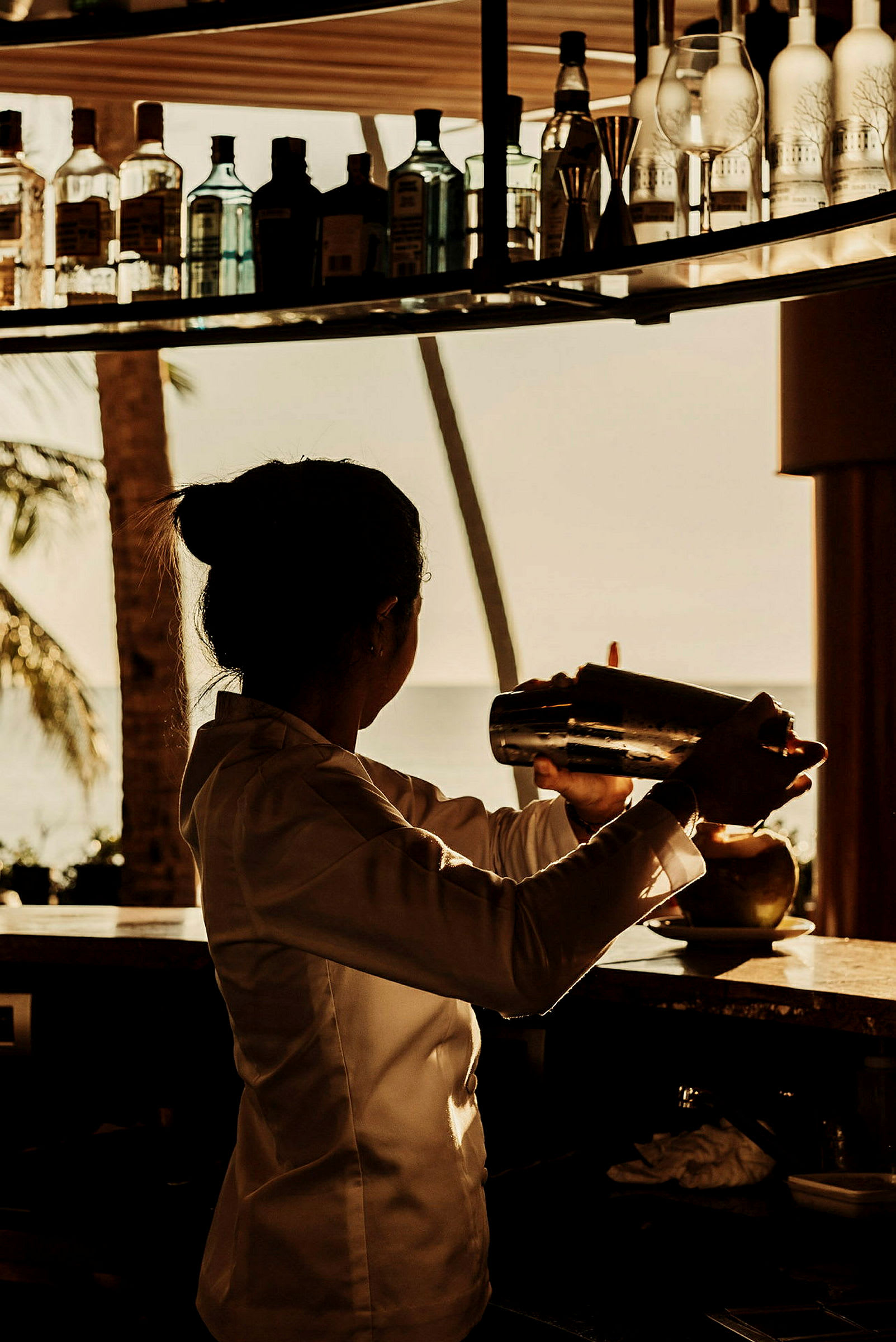 En person i profil håller en cocktailshaker bakom en bardisk. Bakgrunden visar flaskor och ett palmlandskap. (AI-genererad text)