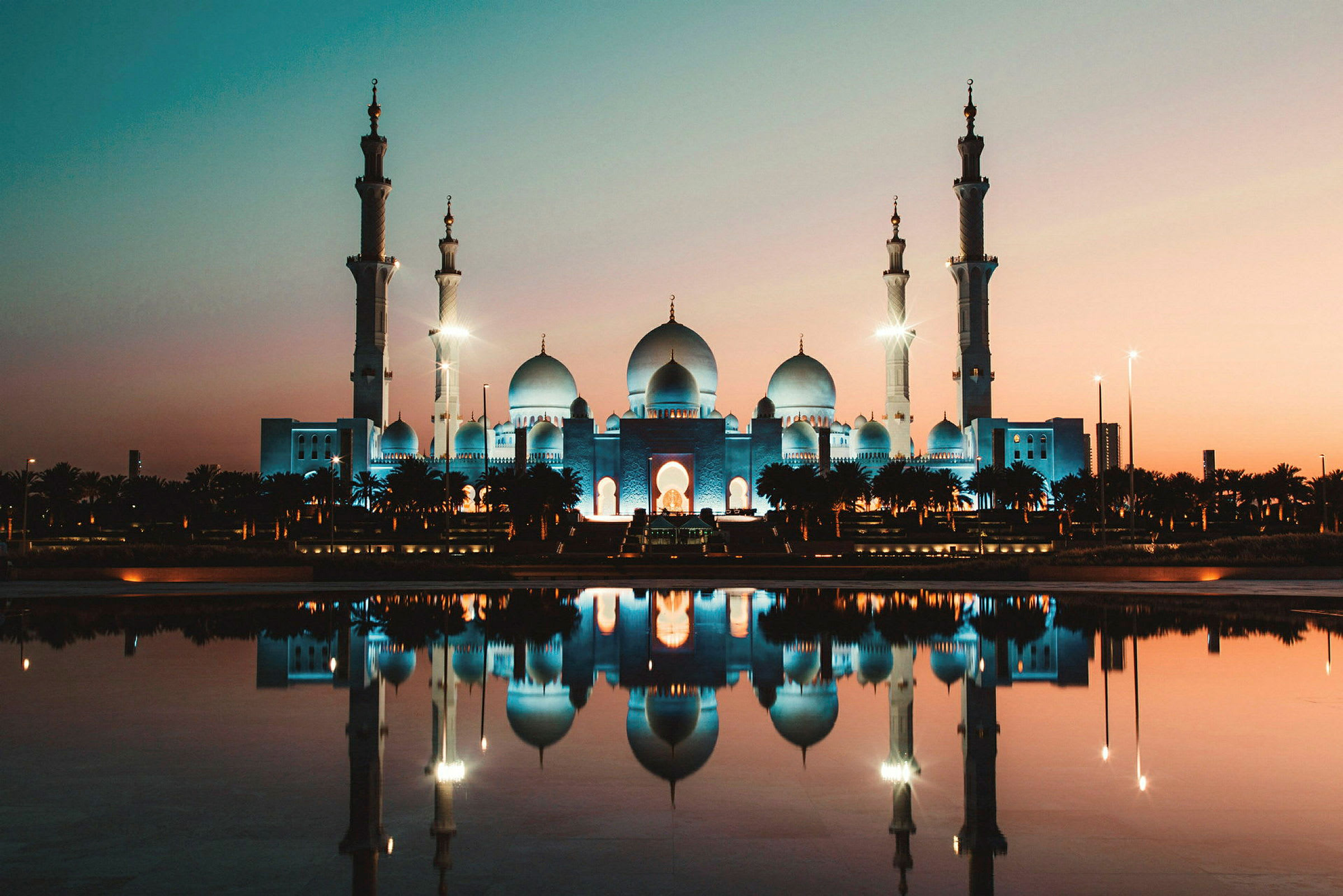 En moské med upplysta minareter och kupoler, reflekteras i vattnet vid solnedgången. (AI-genererad text)