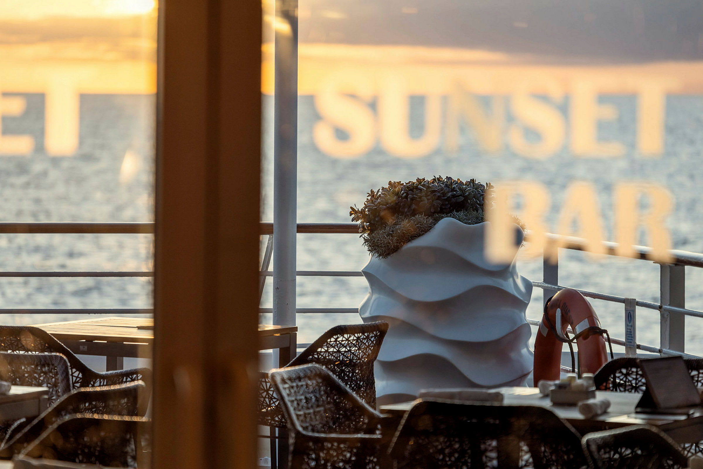 Uteservering med stolar, bord och en växt, havet syns i bakgrunden. (AI-genererad text)