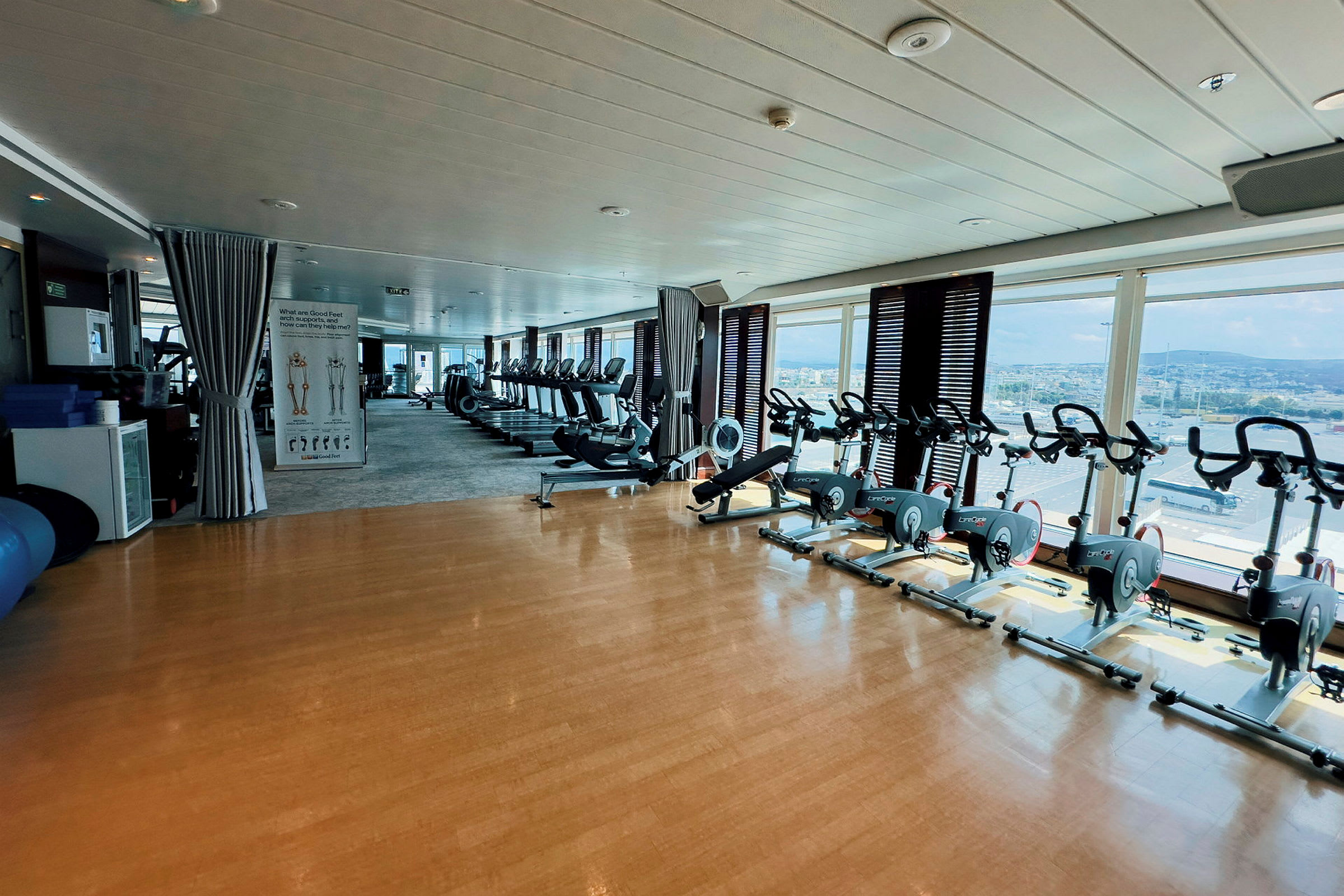 Gym med träningsmaskiner och motionscyklar, stora fönster som ger utsikt över ett städerområdet. (AI-genererad text)
